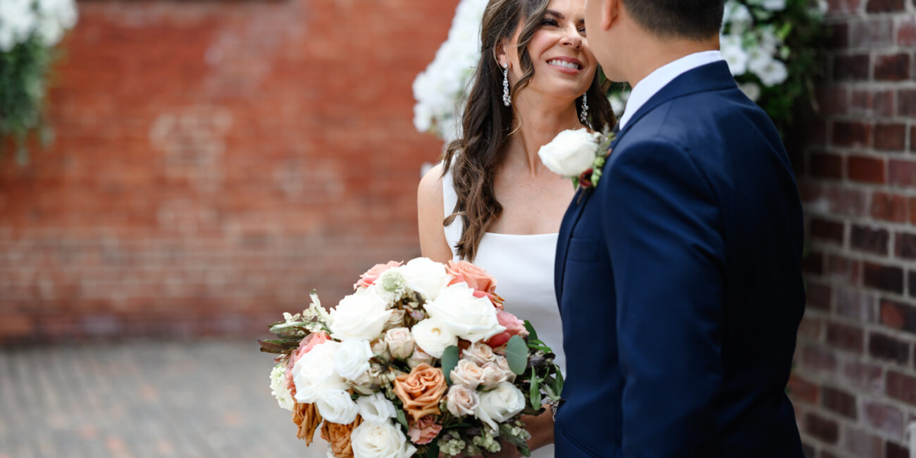 Archeo at The Distillery District: A bride holding a bouquet smiles at a groom in a navy suit, with both standing against a brick wall adorned with white flowers, capturing the enchanting charm of a Distillery District Toronto wedding.