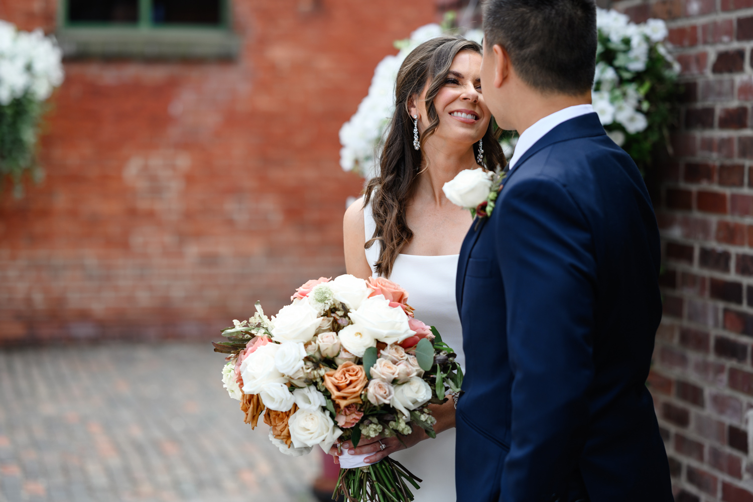 Archeo at The Distillery District: A bride holding a bouquet smiles at a groom in a navy suit, with both standing against a brick wall adorned with white flowers, capturing the enchanting charm of a Distillery District Toronto wedding.