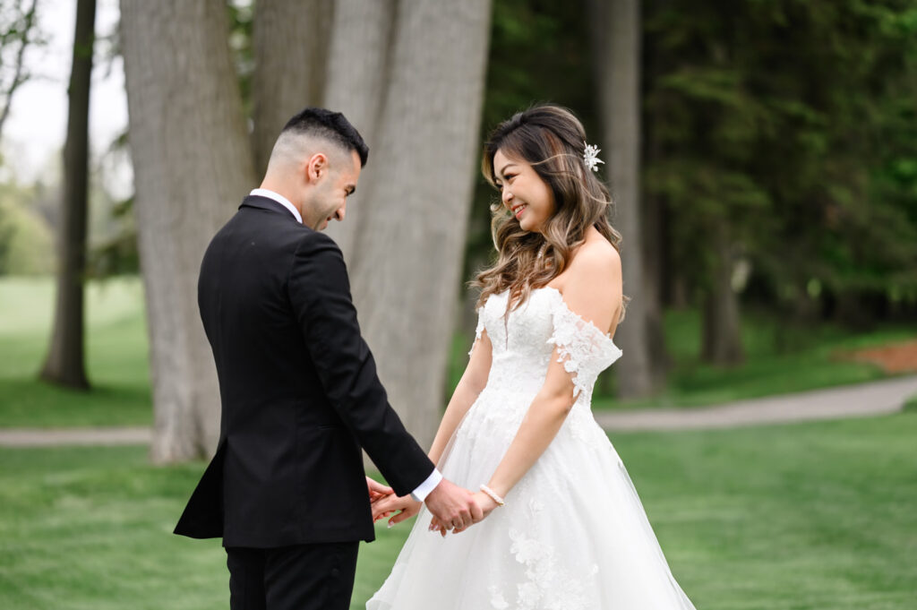 At A Toronto Hunt Golf Club wedding A bride and groom smiling and holding hands in a park, surrounded by trees.