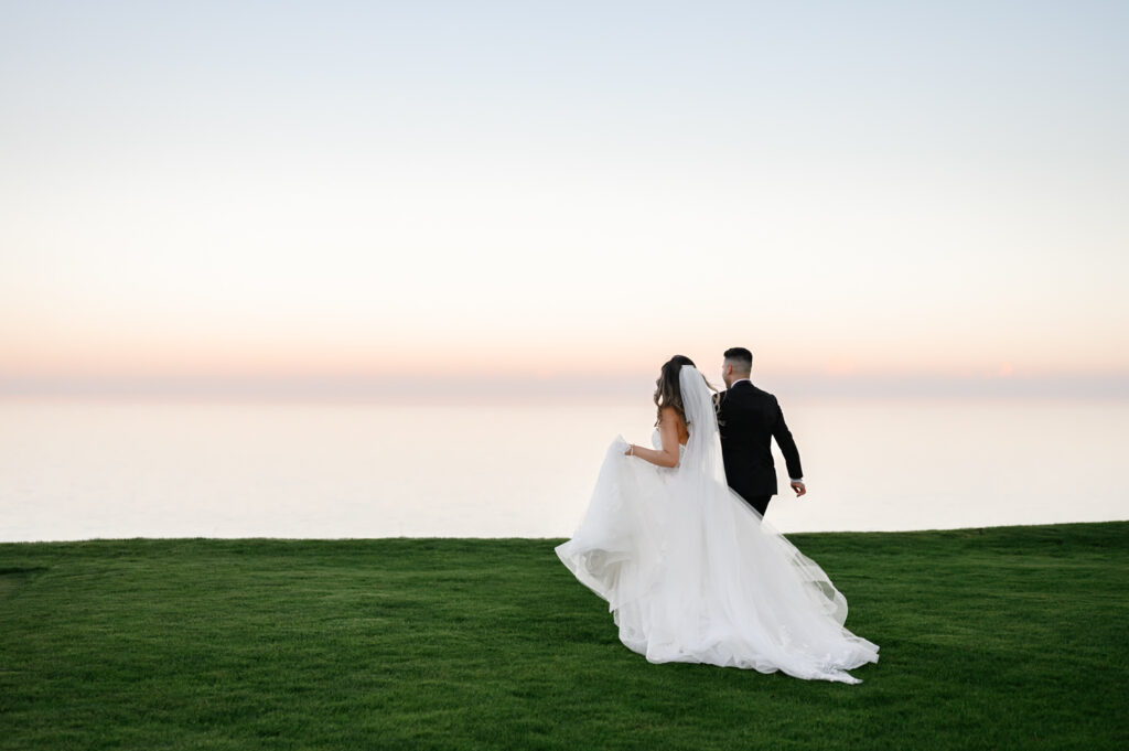 As the sun sets, a bride and groom stroll across a grassy field at The Toronto Hunt Wedding, overlooking the calm sea.