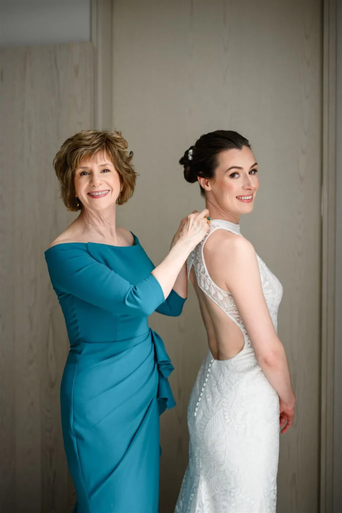 A woman in a teal dress fastens the back of a bride’s lace wedding gown at Pearle Hotel Weddings. Both are smiling, standing indoors against a light wood background.