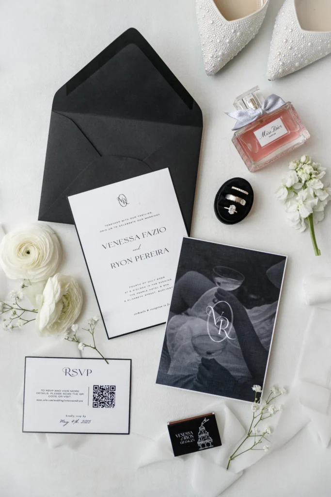 Flat lay of wedding invitation suite with RSVP card, black envelope, rings, white flowers, perfume, and white shoes on a white surface—perfect inspiration for your Pearle Hotel celebration.