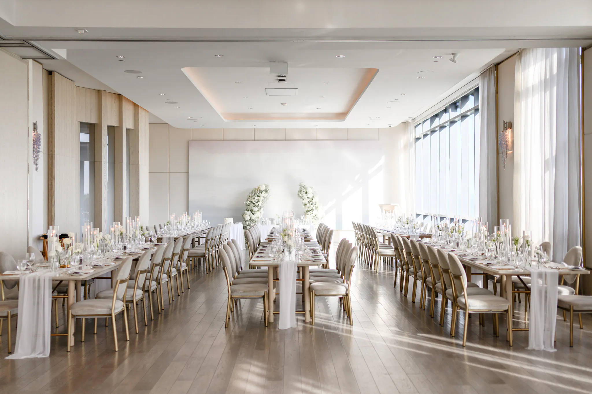 A bright, elegant banquet hall at the Pearle Hotel is set for an event with long tables, gold chairs, white flowers, and candles, with natural light streaming through large windows.