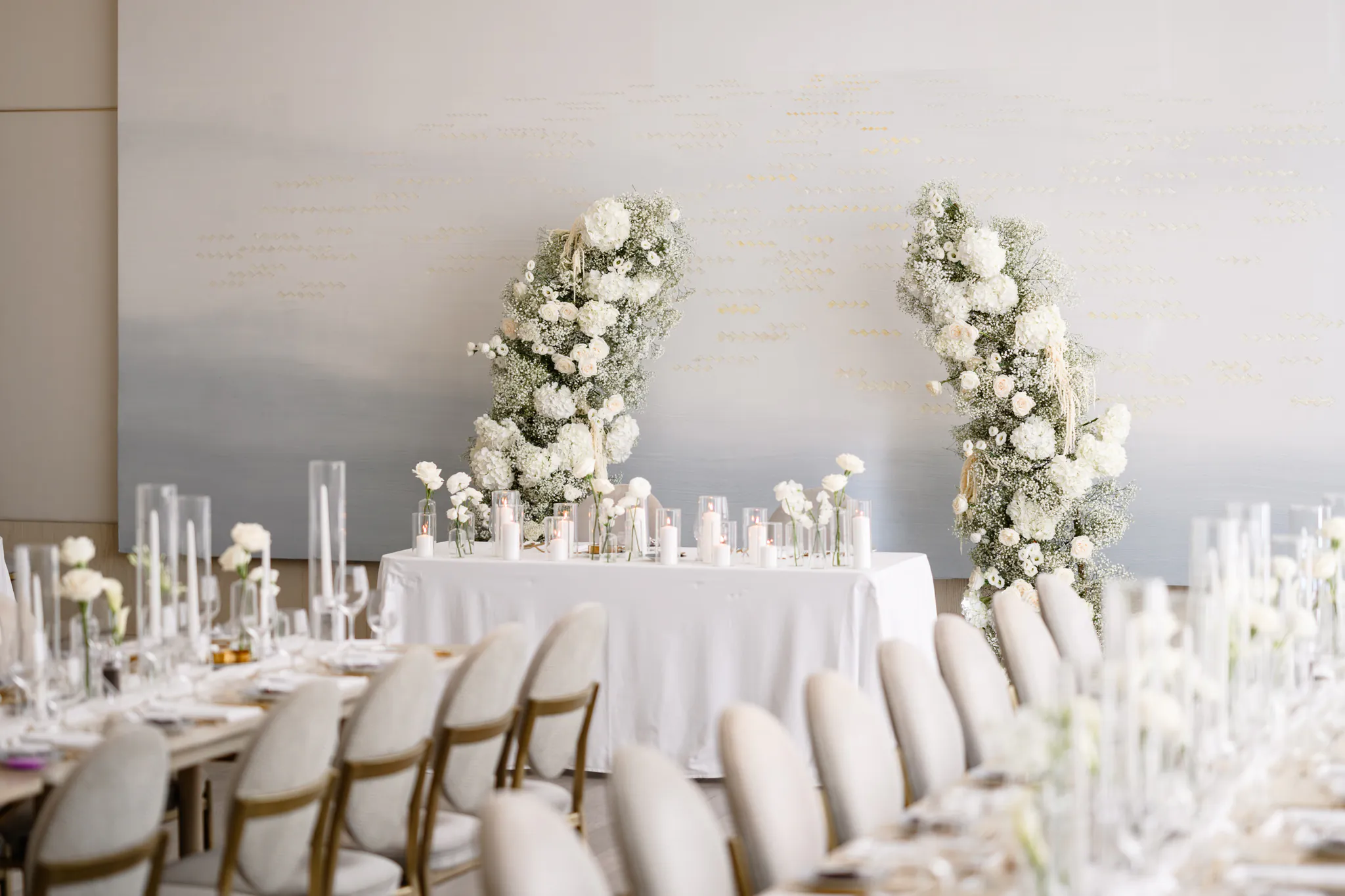 A wedding reception setup at the Pearle Hotel with a long table, white chairs, floral centerpieces, and two large white flower arrangements behind a head table.