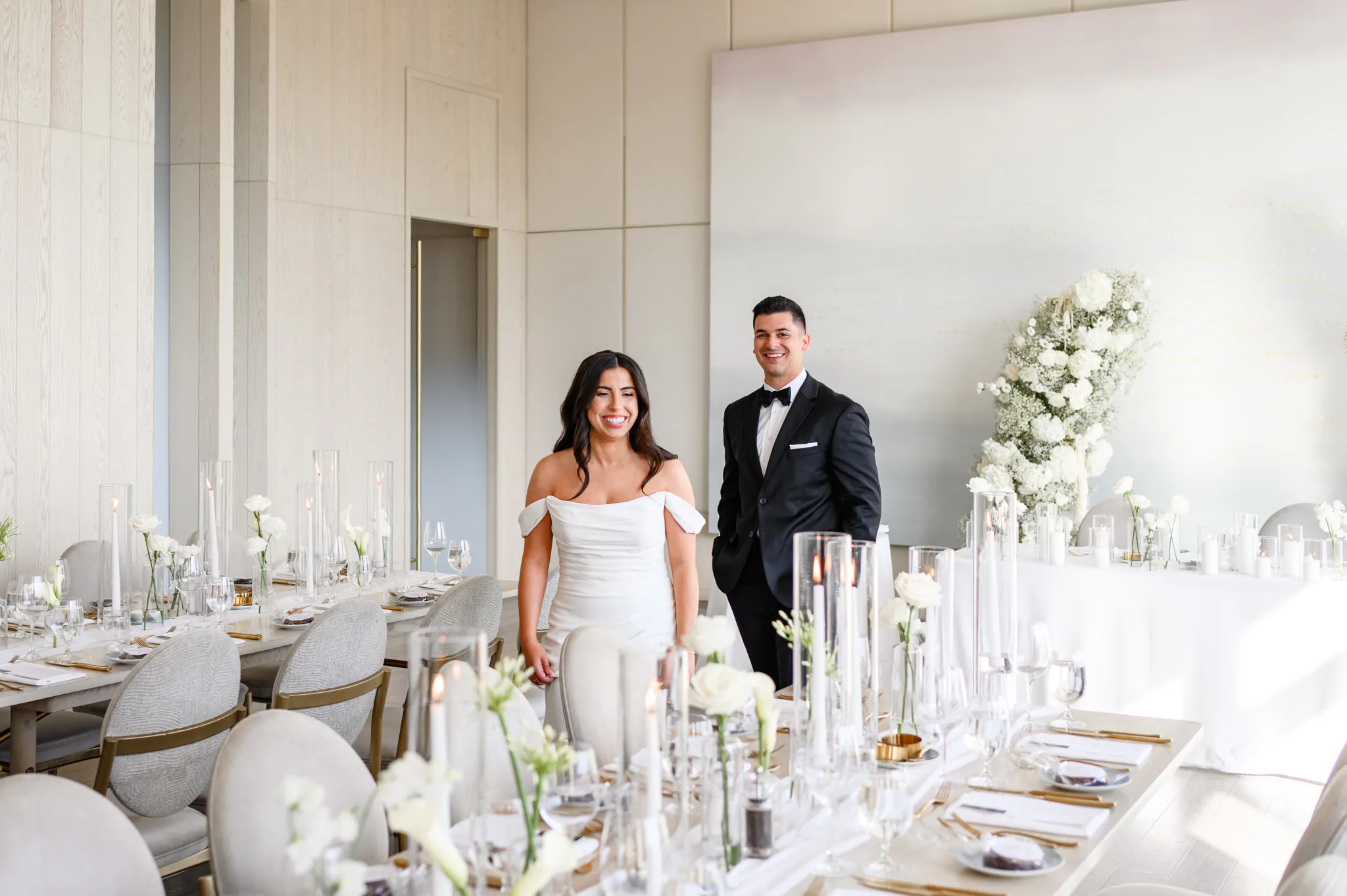 A bride and groom stand smiling in the elegantly decorated Pearle Hotel reception room, adorned with white flowers, candles, and formal table settings.