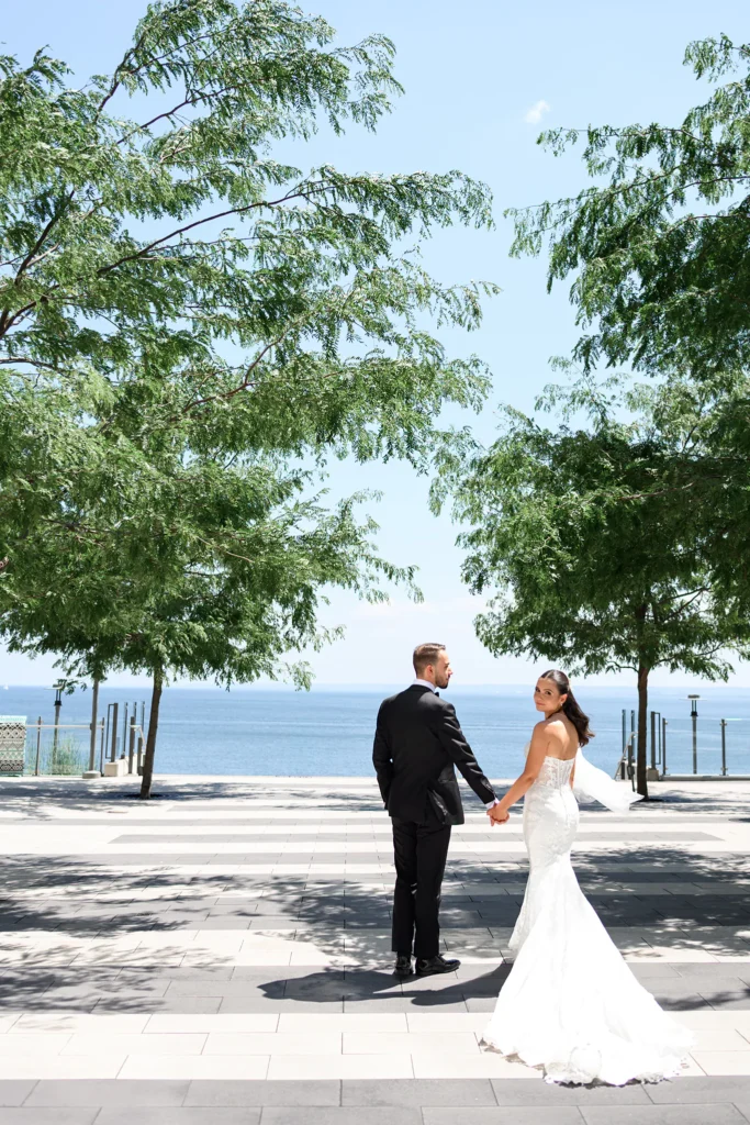 A bride in a white gown and a groom in a black suit hold hands and look back while standing on a sunlit waterfront promenade lined with green trees.