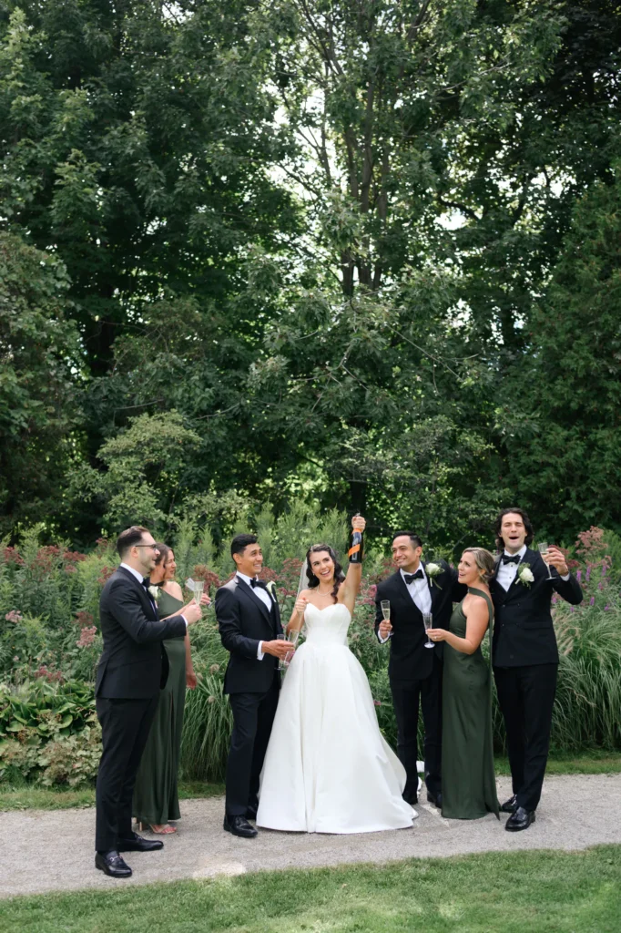 A bride and groom stand outdoors at Paletta Mansion with four members of their wedding party, all raising glasses and smiling in formal attire with lush greenery in the background.