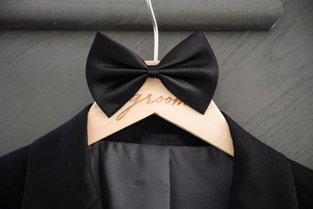 A black tuxedo jacket on a wooden hanger labeled "groom," with a black bow tie attached—ready for an elegant Pearle Hotel Wedding.