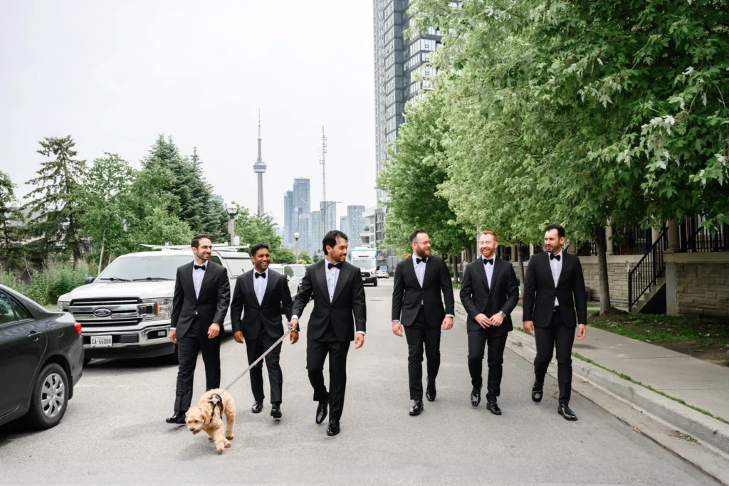 Six men in black tuxedos walk down a city street with a dog on a leash; tall buildings and the CN Tower set the backdrop for The Quay Wedding.