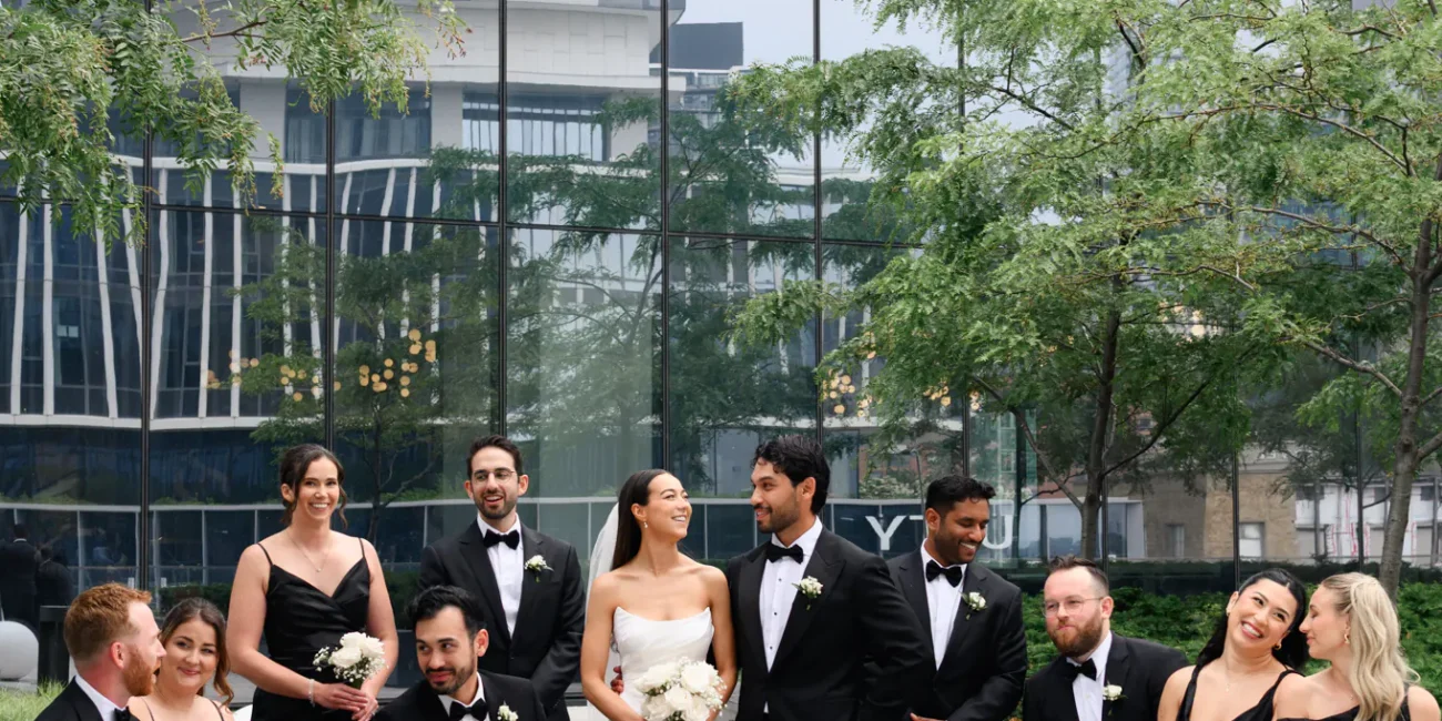 A wedding party in formal attire poses outdoors at The Quay Wedding, in front of a modern glass building, with the bride and groom standing at the center holding bouquets.