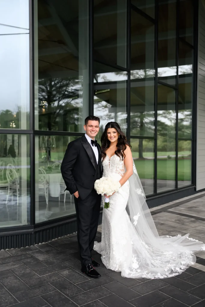 A bride in a white wedding dress and a groom in a black tuxedo stand together outside the large glass windows at their Three Feathers Terrace Wedding.
