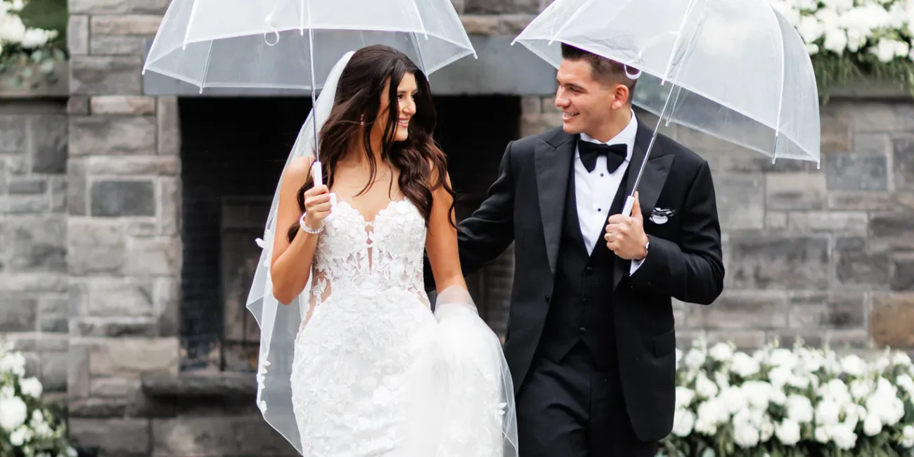 A bride and groom in formal attire walk together outdoors, each holding a clear umbrella, in front of a stone backdrop with white flowers at their Three Feathers Terrace Wedding.