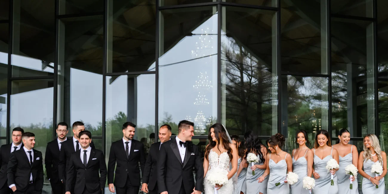 A wedding party poses outside Three Feathers Terrace Wedding venue in front of large glass windows, with groomsmen in black suits and bridesmaids in light gray dresses holding white flowers.