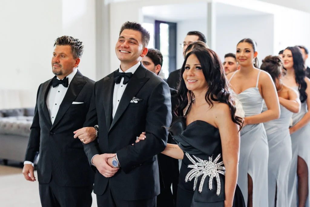 A group of people dressed formally at a Three Feathers Terrace Wedding, with the man in the center smiling and linking arms with a woman, while others stand behind them in gray dresses and tuxedos.