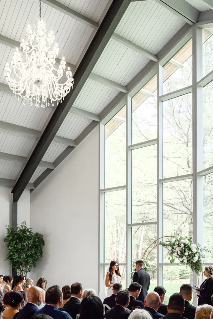 A bride and groom stand at the altar in a bright chapel with large windows, exchanging vows before seated guests at their elegant Three Feathers Terrace Wedding.