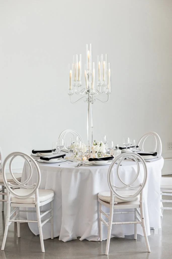 Round white table set with black napkins, glassware, and a tall silver candelabra centerpiece, surrounded by four white chairs—an elegant setting inspired by a Three Feathers Terrace Wedding against a plain white background.