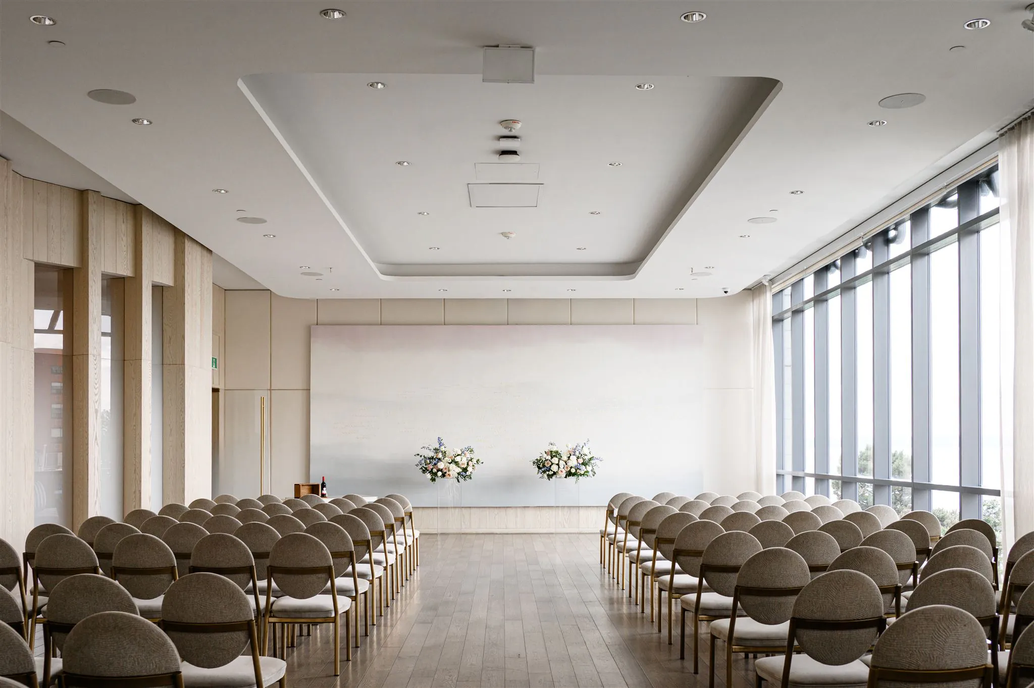 A modern, light-filled ceremony room with rows of beige chairs facing a simple altar adorned with two floral arrangements. Large windows line one side of the room.
