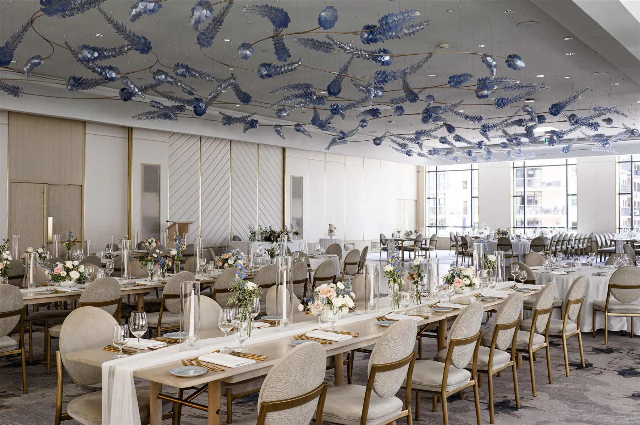Elegant banquet hall with long dining tables, floral centerpieces, neutral-toned chairs, and blue decorative ceiling accents, set for a formal event.