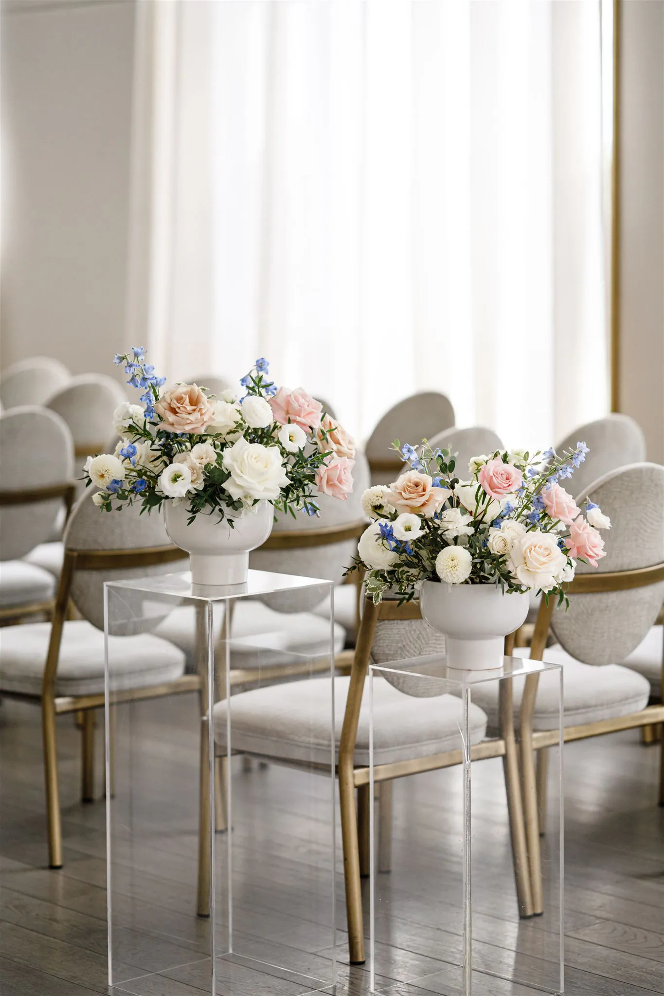 Two white vases with pastel flower arrangements sit on clear stands in front of rows of beige and gold chairs in a bright room.