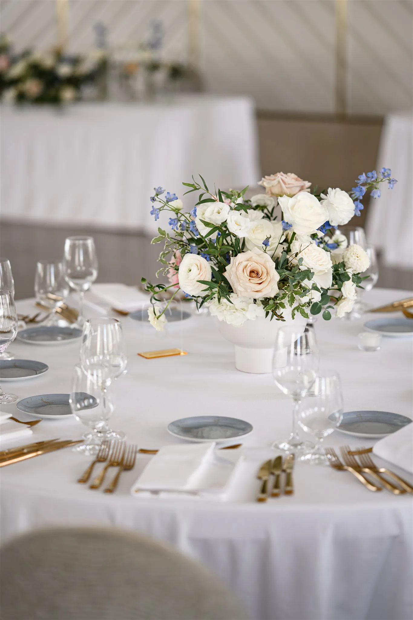 A round dining table set with white linens, gold cutlery, glassware, and a centerpiece of white and pale pink flowers with greenery.