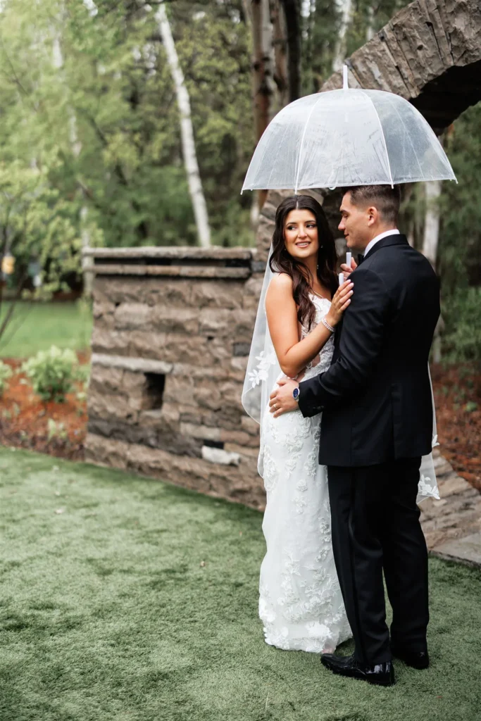 A bride and groom stand close together under a transparent umbrella on a grassy area at Three Feathers Terrace, with trees and stone structures in the background.