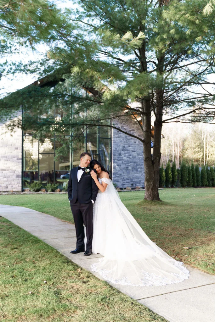 A bride in a white gown and a groom in a black tuxedo stand together on a sidewalk outside the modern glass building of Three Feathers Terrace, surrounded by green trees and grass—perfect for memorable three feathers terrace photos.