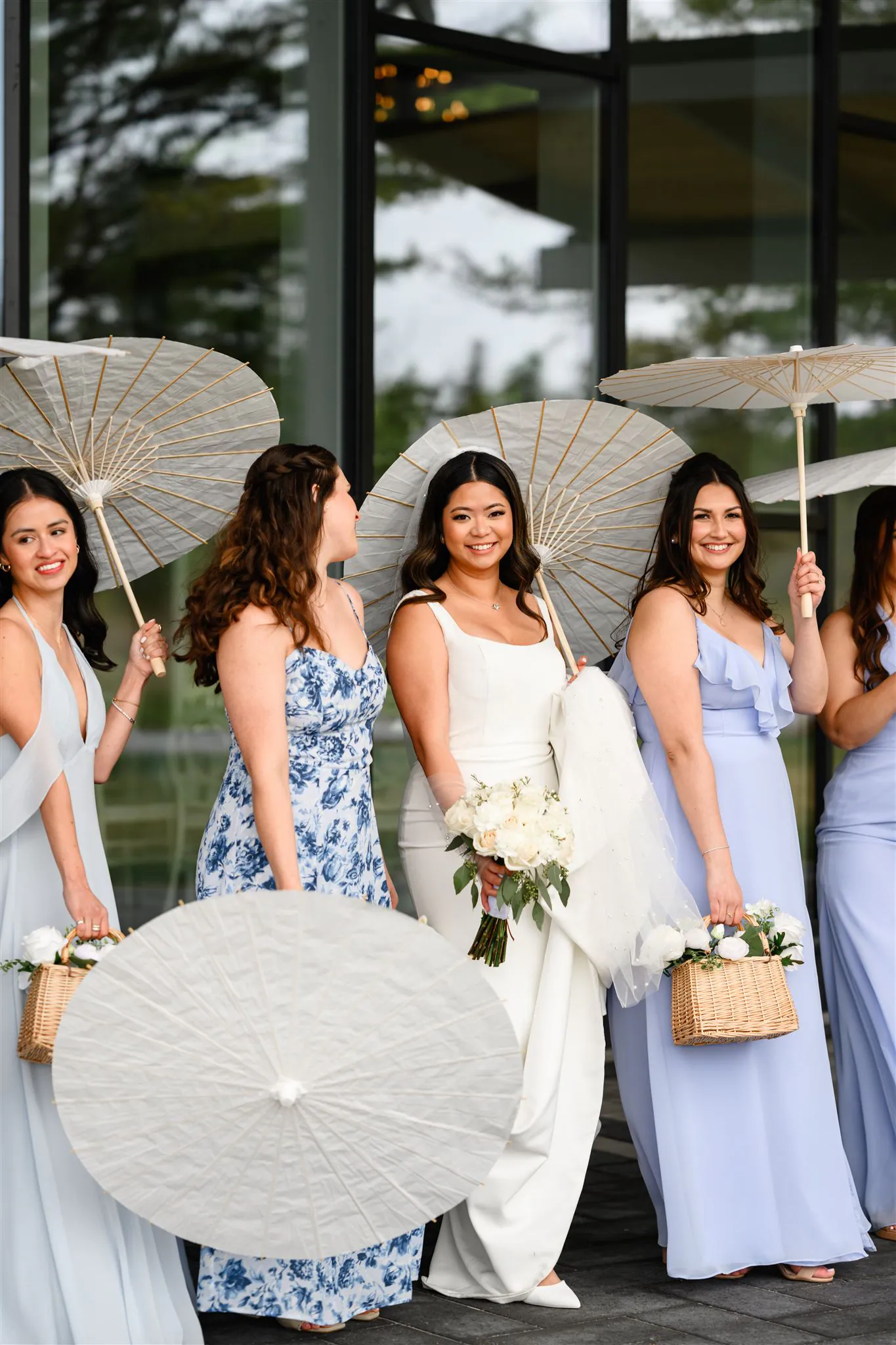 A bride in a white dress stands with her bridesmaids holding white parasols and small bouquets, in front of the stunning glass facade at Three Feathers Terrace. Perfect for capturing elegant three feathers terrace photos on your special day.