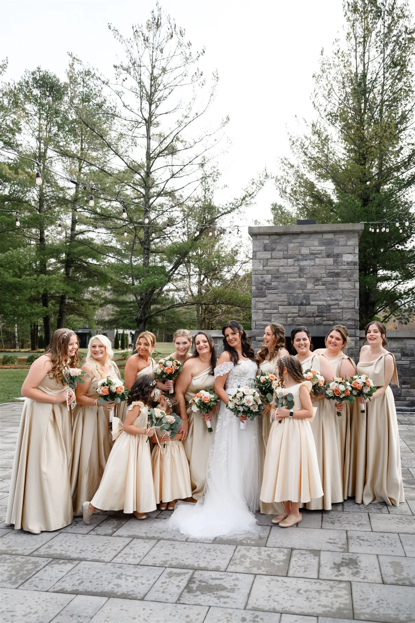 A bride in a white gown stands with twelve bridesmaids in matching champagne dresses, all holding bouquets, outdoors on a stone patio at Three Feathers Terrace with tall trees in the background—perfect for stunning three feathers terrace photos.