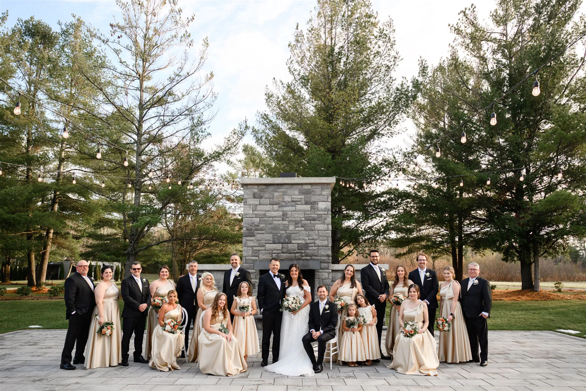 A wedding party poses outdoors on a stone patio at Three Feathers Terrace, in front of a stone structure with trees in the background. The group includes the bride, groom, bridesmaids, and groomsmen. Contact for Three Feathers Terrace wedding cost details.