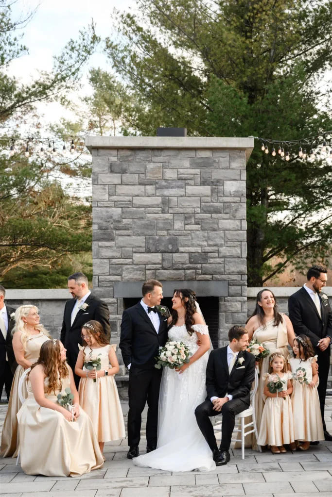 A bride and groom stand together in front of an outdoor stone fireplace at Three Feathers Terrace, surrounded by bridesmaids in gold dresses and groomsmen in black suits.