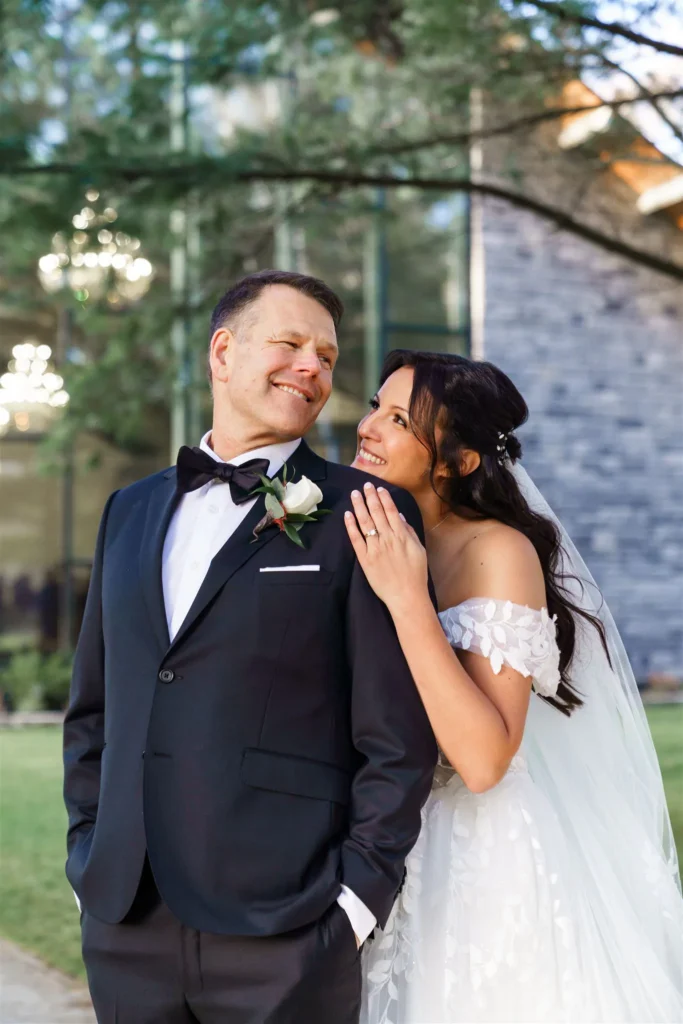 A bride in a white wedding dress smiles at and leans on a groom in a black tuxedo outdoors at Three Feathers Terrace, with lush greenery and a building in the background, capturing the magic found in Three Feathers Terrace photos.