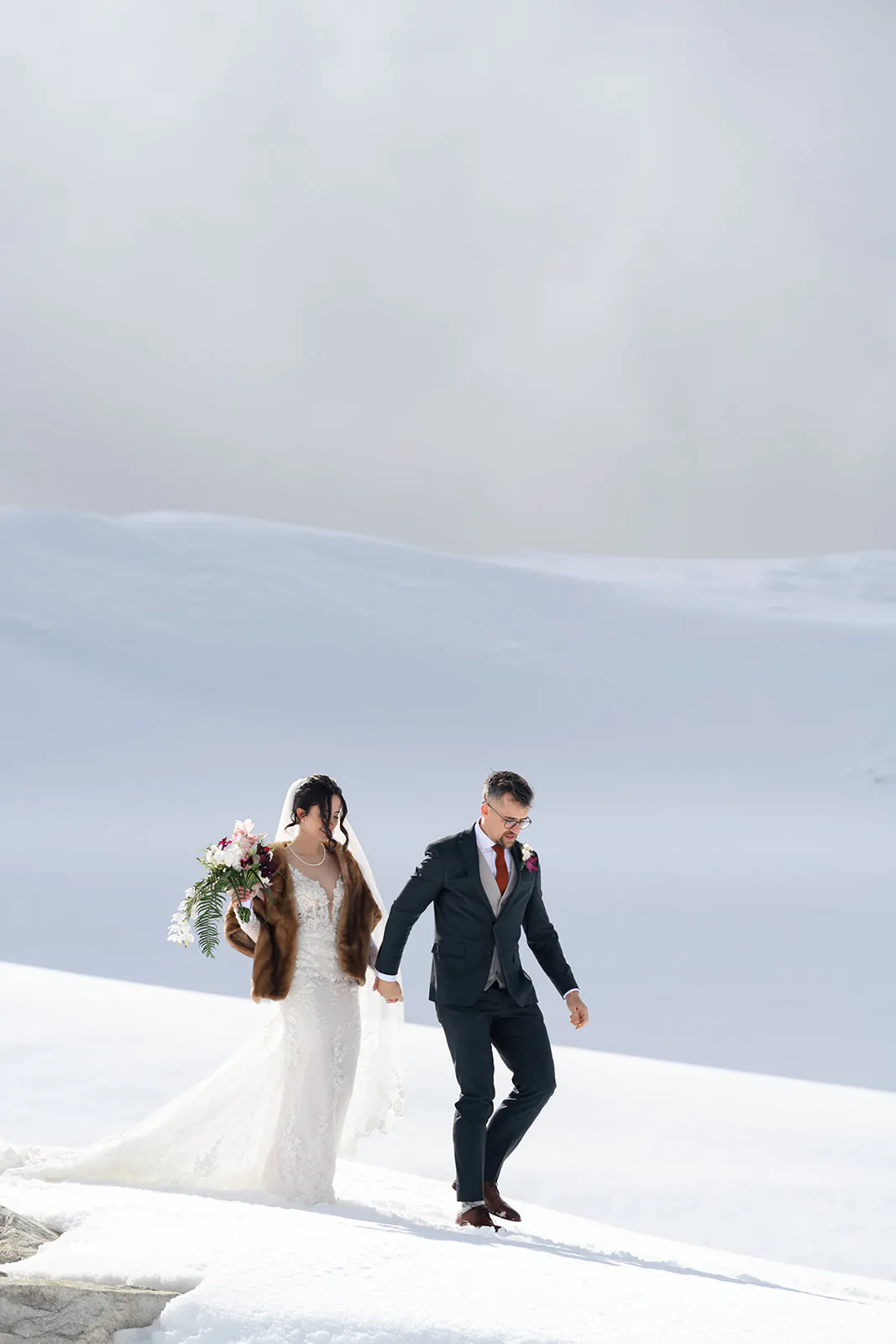 A bride in a white dress and brown shawl holds a bouquet while walking beside her groom in a suit across a snowy landscape, capturing the magic of a Whistler elopement wedding.