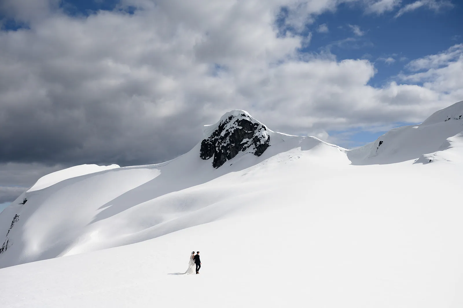 A couple stands on a snowy mountain slope under a partly cloudy sky, with a rugged peak in the background—perfect for a Whistle bear wedding amid nature’s grandeur.