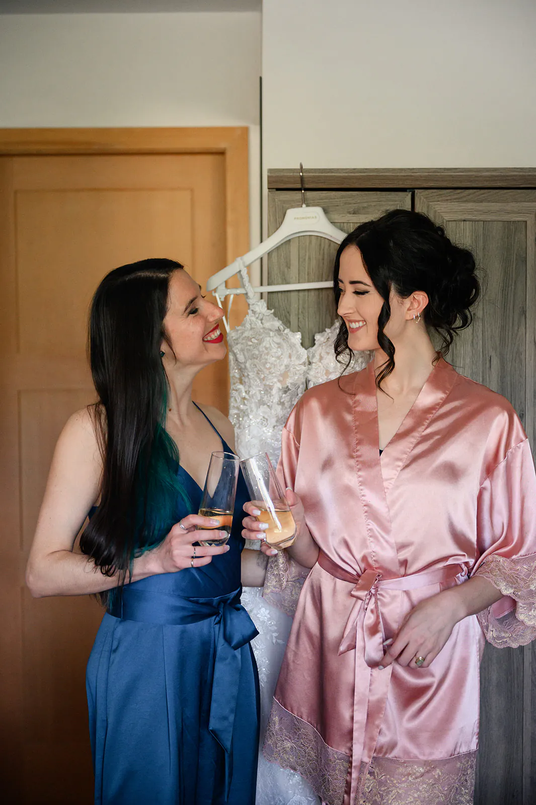 Two women smile at each other and hold drinks; one wears a blue dress, the other a pink robe with a white wedding dress hanging in the background.