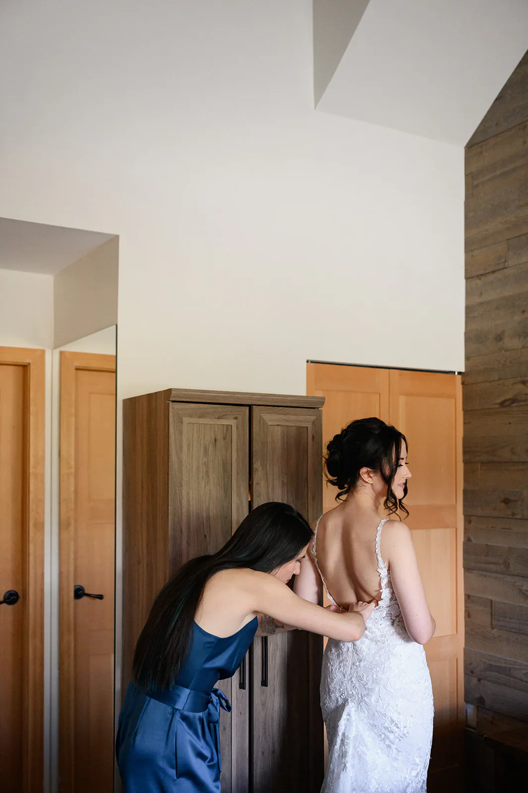 A woman in a blue dress fastens the back of a bride’s white gown in a room with wooden doors and cabinets.