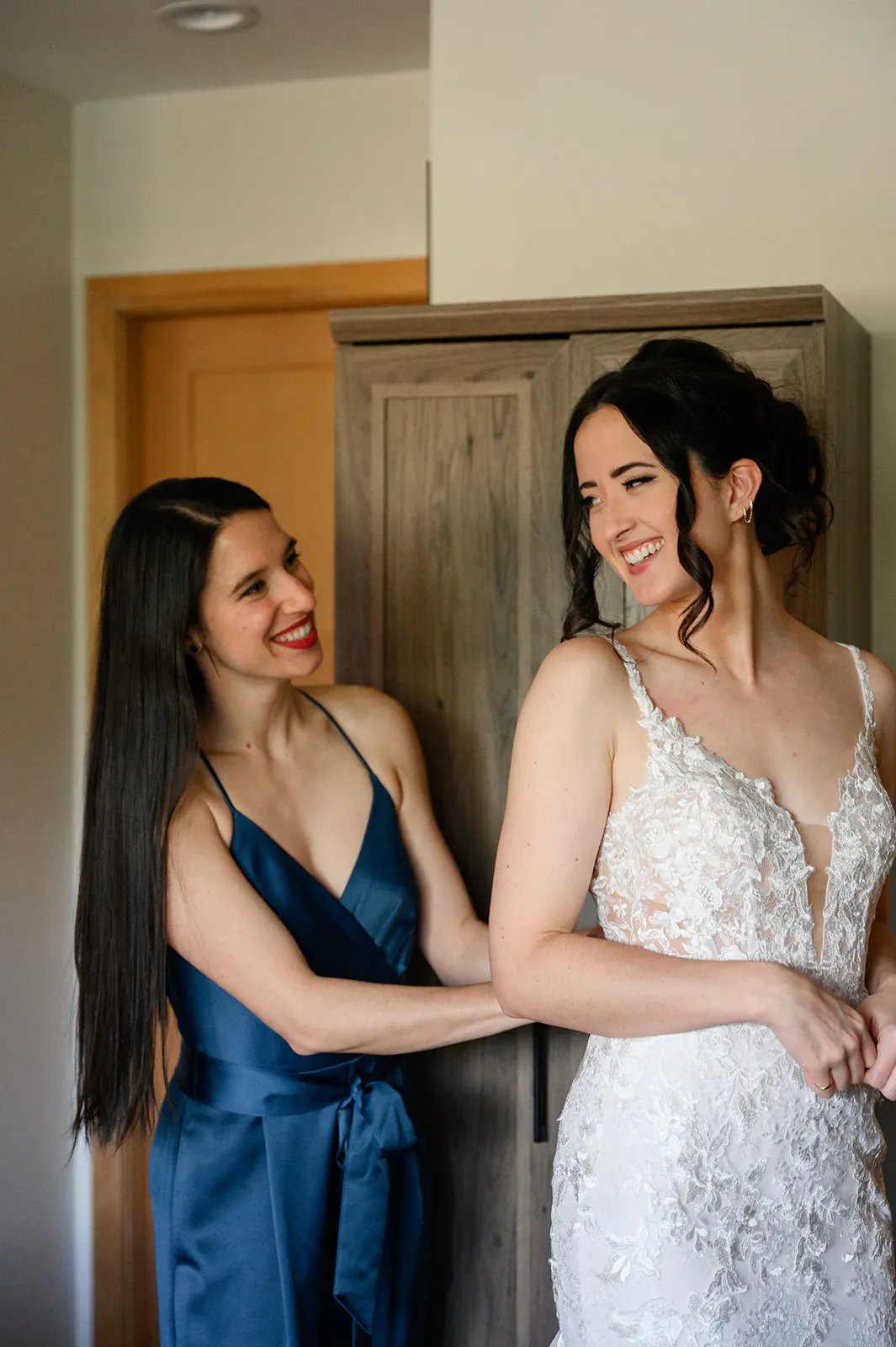 Two women, one in a blue dress and the other in a white lace wedding gown, smile at each other while standing indoors near a wooden wardrobe.