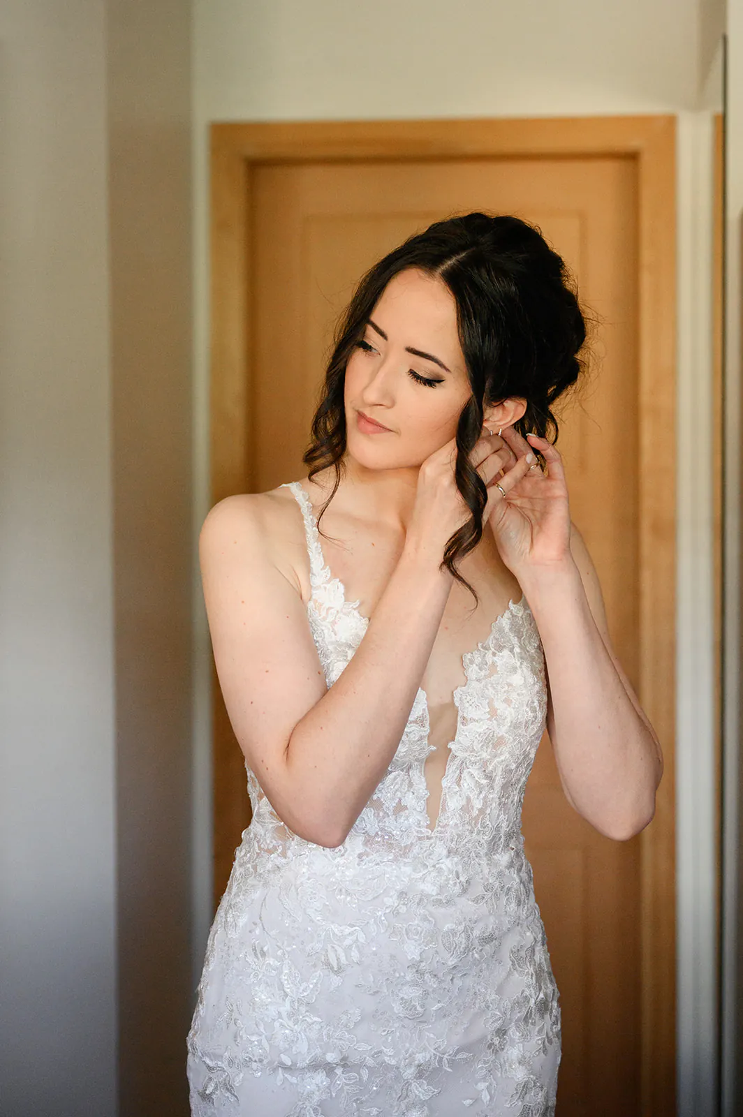 A woman in a white lace wedding dress adjusts an earring while standing indoors near a wooden door.