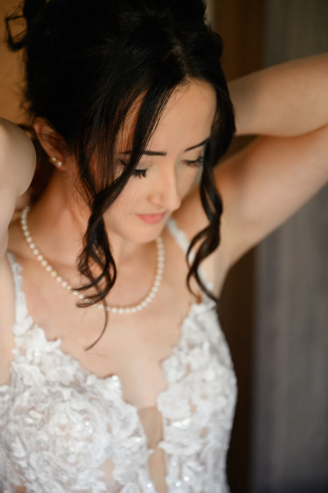 Woman in a white lace wedding dress adjusts her necklace, looking down with dark hair styled in loose curls.