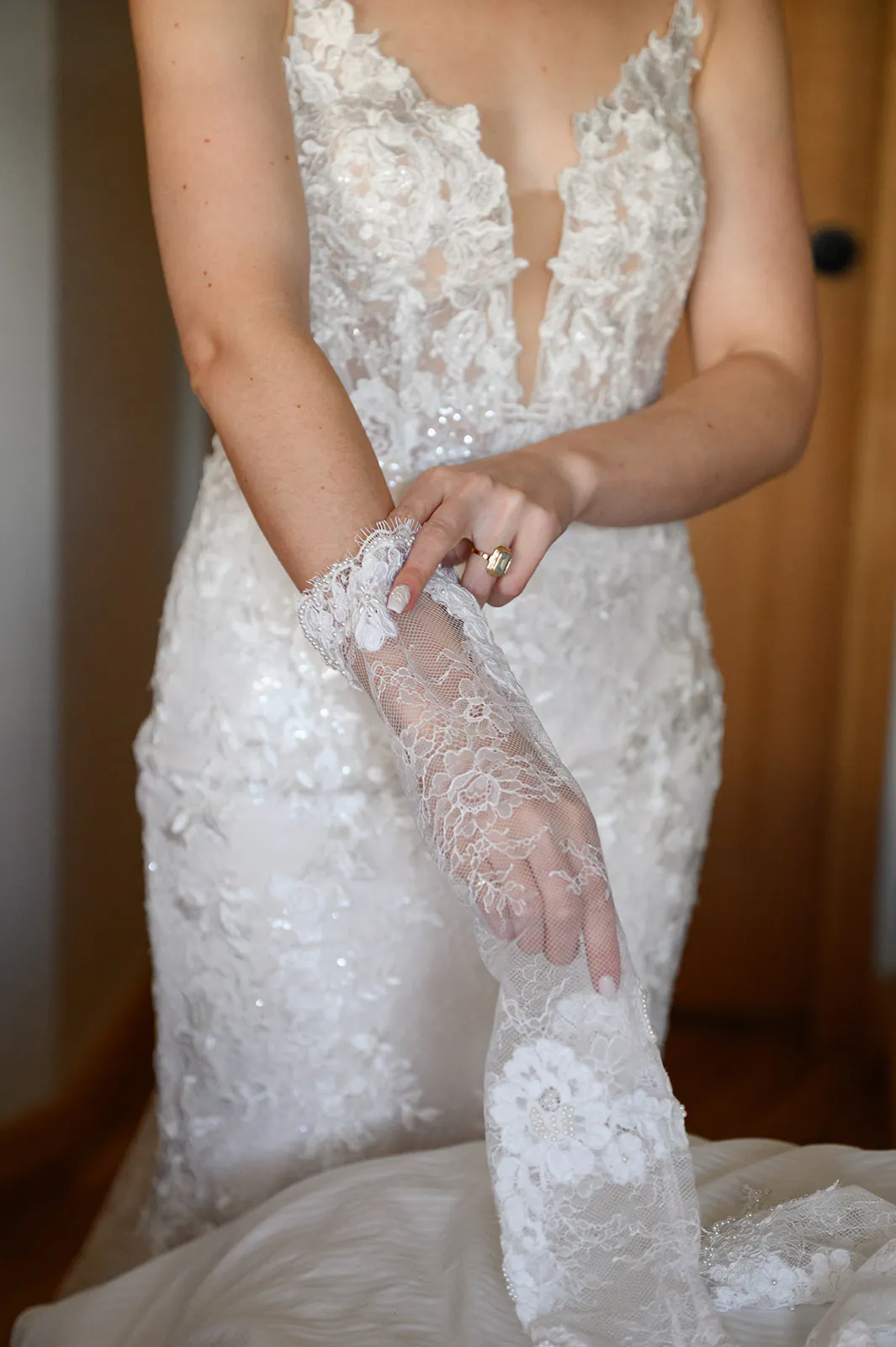 A person in a white, floral lace wedding dress puts on a matching lace glove, standing indoors near a wooden door.