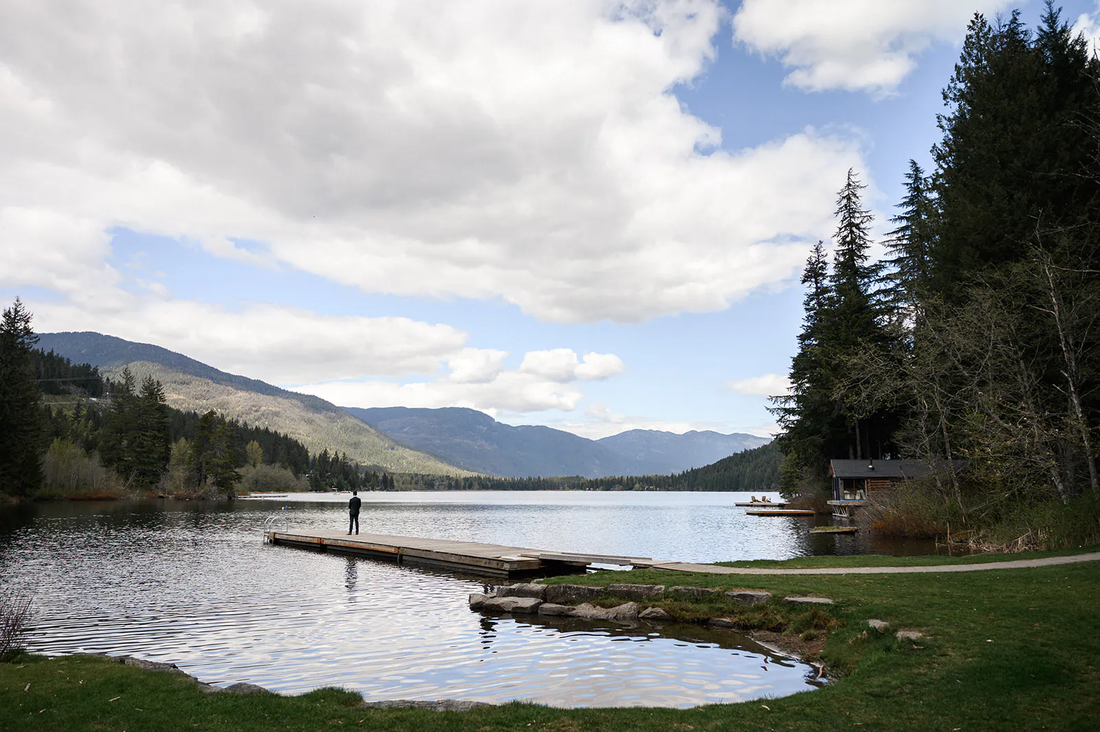A person stands on a wooden dock extending into a calm lake, surrounded by trees and mountains under a partly cloudy sky.