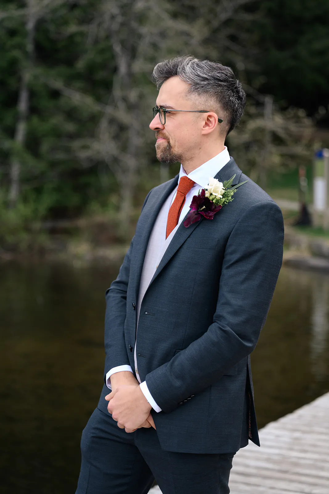 Man in a dark suit with a red tie and boutonnière stands on a wooden dock near water, looking to the side with trees in the background.