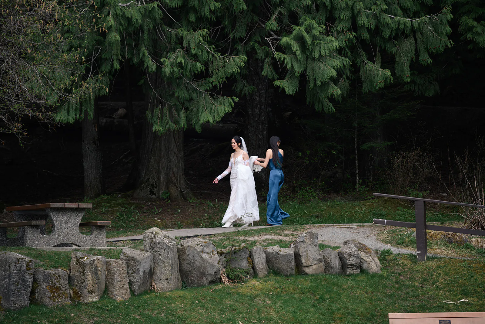 A bride in a white dress and another woman in a blue dress walk together on a path in a wooded park area.