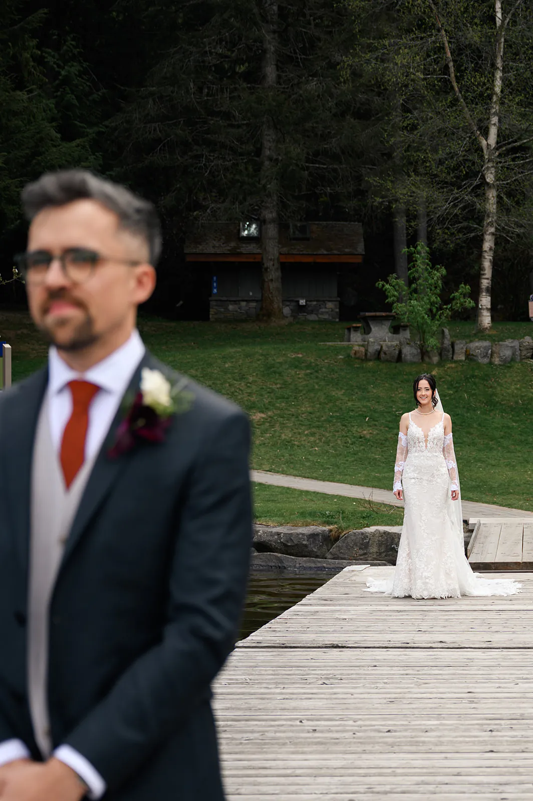 A bride in a white wedding dress walks toward a groom in a suit, standing on a wooden dock by a grassy area with trees.