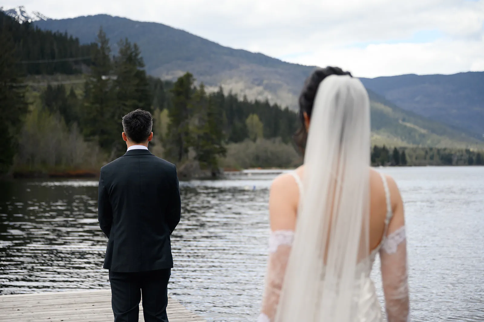 A bride in a white dress approaches a groom in a dark suit standing on a dock by a lake, with mountains and trees in the background.