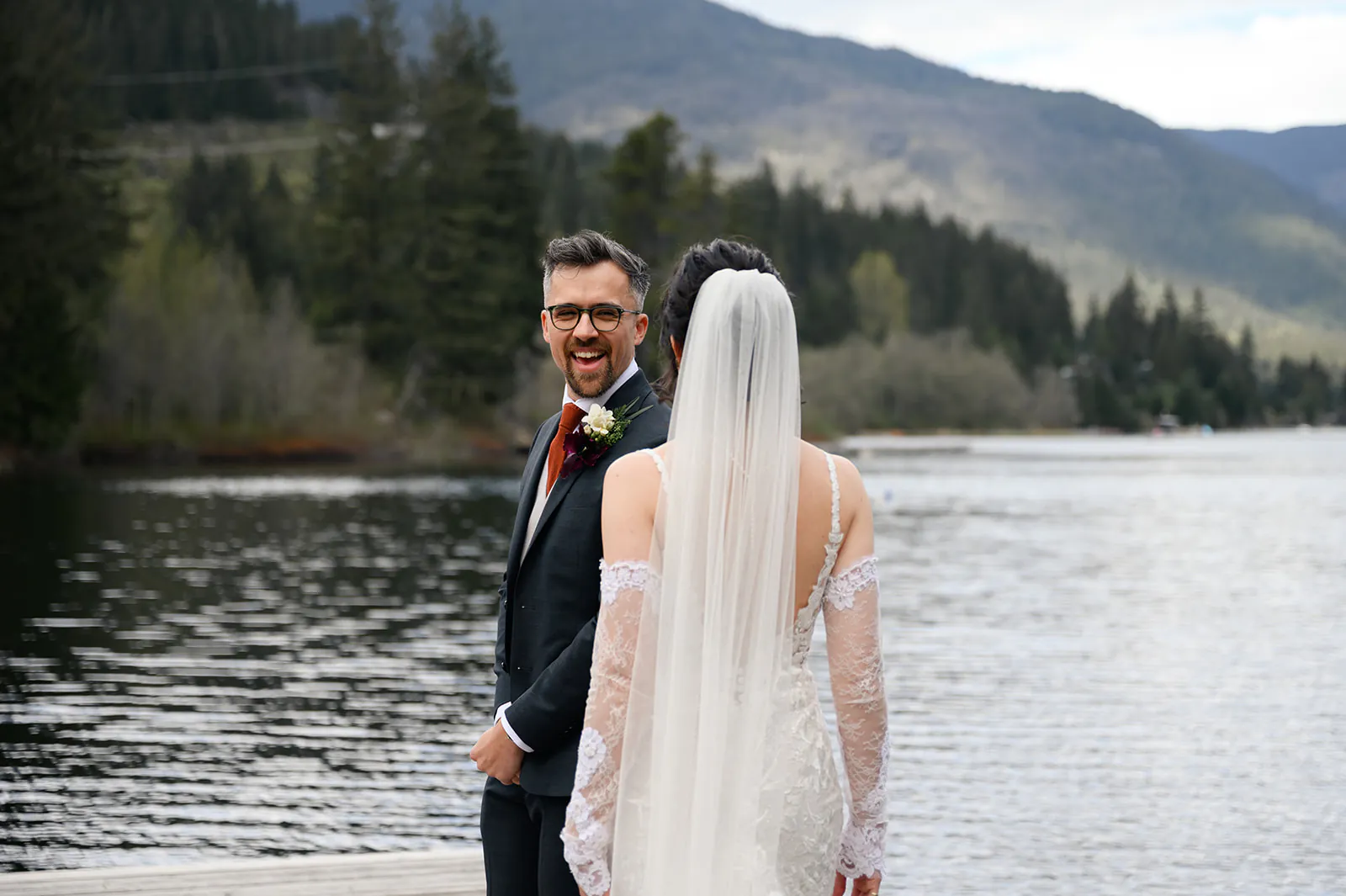 A groom in a suit smiles at a bride in a veil and lace dress as they stand on a lakeside dock with mountains and trees in the background.