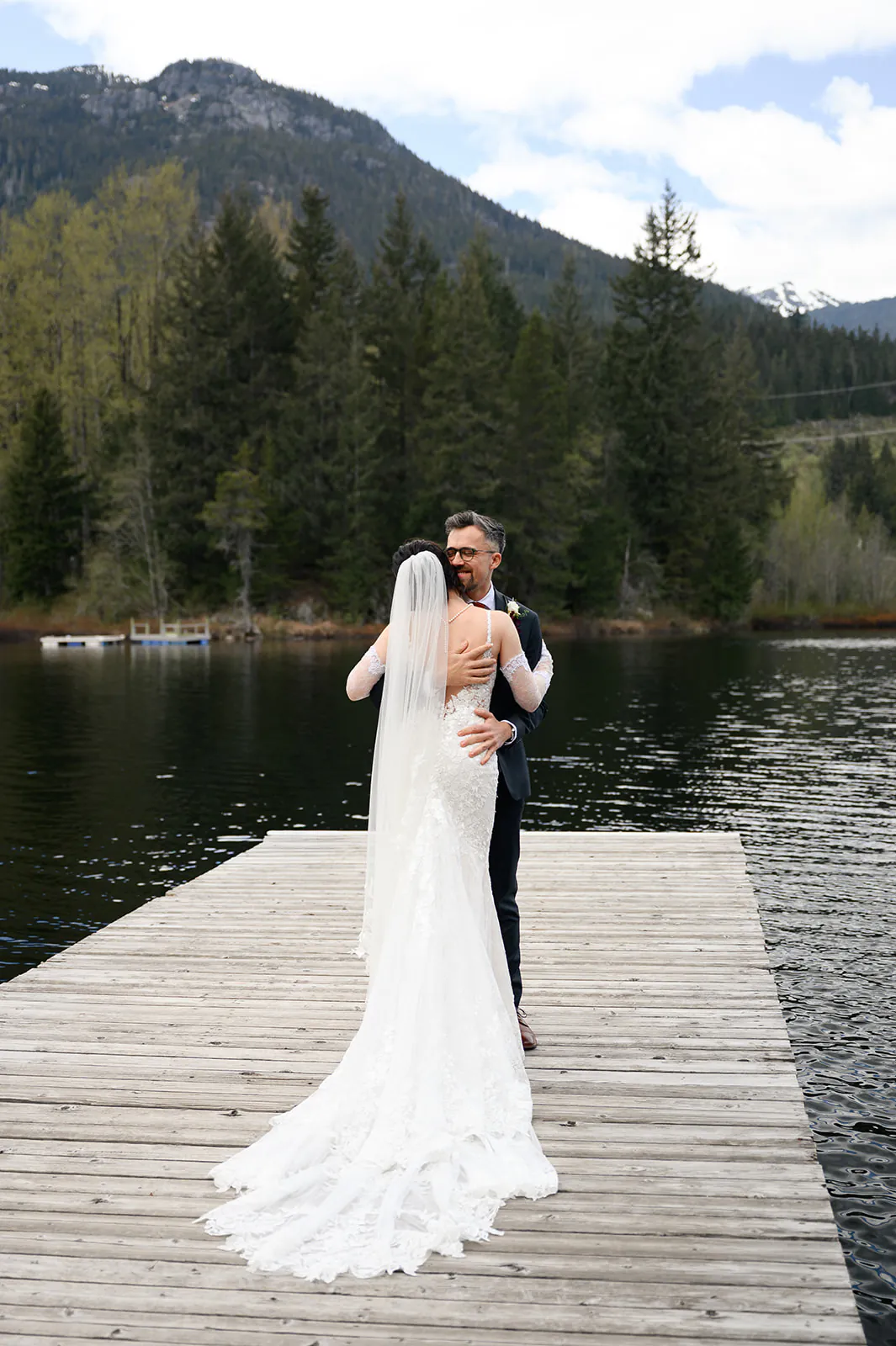 A bride and groom embrace on a wooden dock by a lake, surrounded by trees and mountains under a partly cloudy sky.