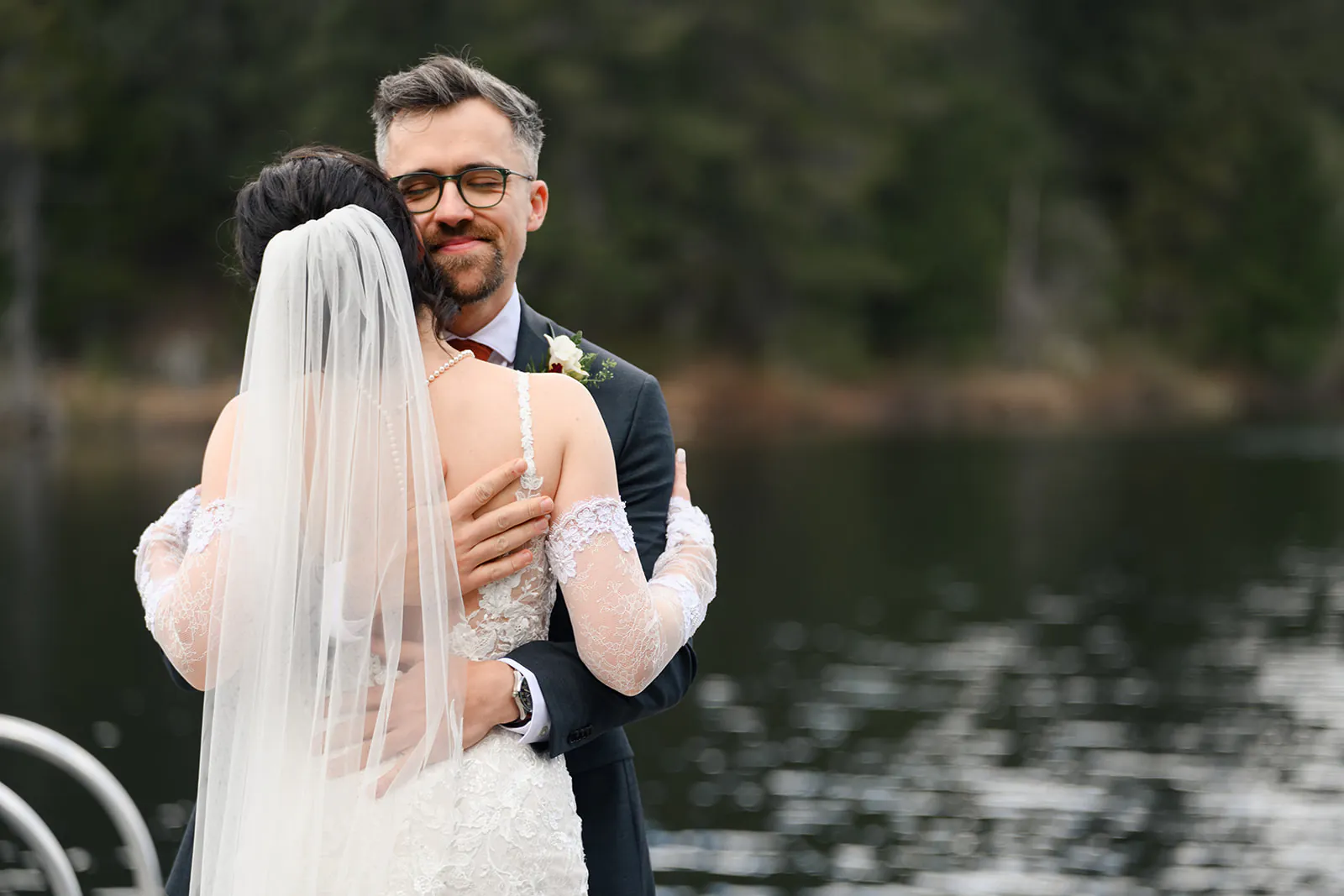 A newlywed couple embraces by a lake, with the groom in a suit and the bride in a lace dress and veil, surrounded by trees in the background.