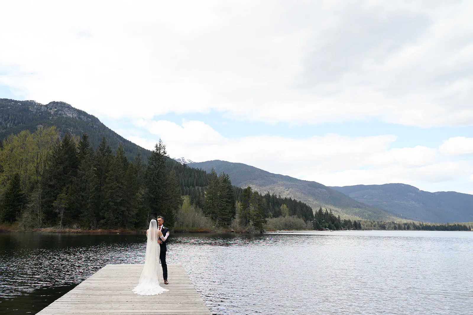 A bride and groom stand together on a wooden dock extending into a lake, surrounded by trees and mountains under a partly cloudy sky.