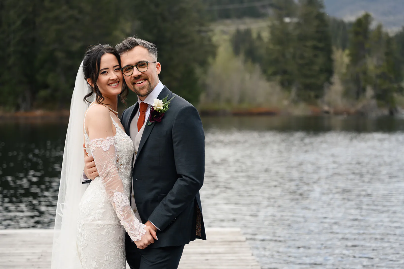 A bride and groom stand on a wooden dock by a lake, smiling and holding hands, with trees and mountains in the background.