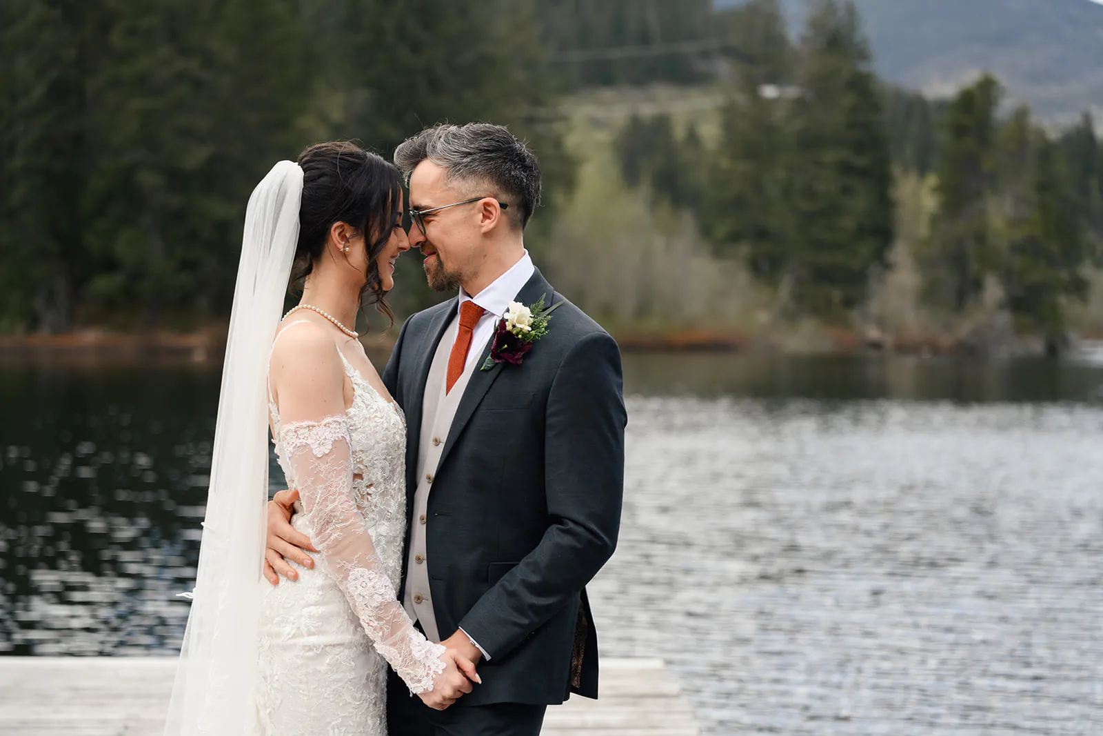 A bride and groom stand close together on a dock by a lake, holding hands and smiling at each other, with trees and mountains in the background.